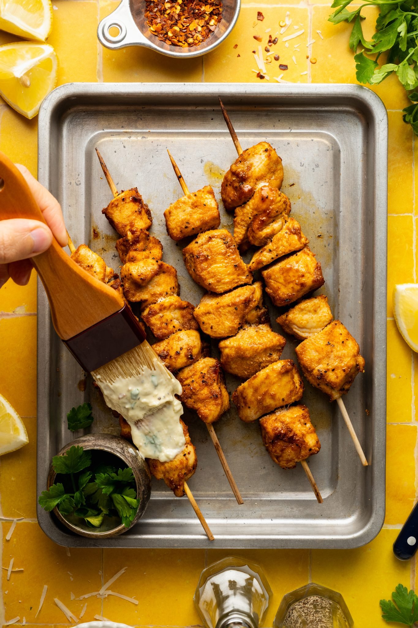 Close-up of golden brown chicken skewers on a sheet pan being brushed with creamy garlic parmesan aioli, surrounded by lemon wedges, parsley, and red pepper flakes.