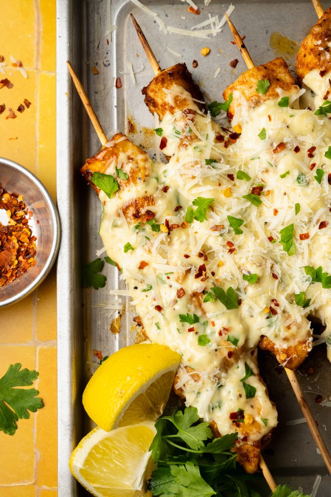 Chicken skewers fully brushed with garlic parmesan aioli and topped with shredded cheese, fresh parsley, and chili flakes on a metal sheet pan.