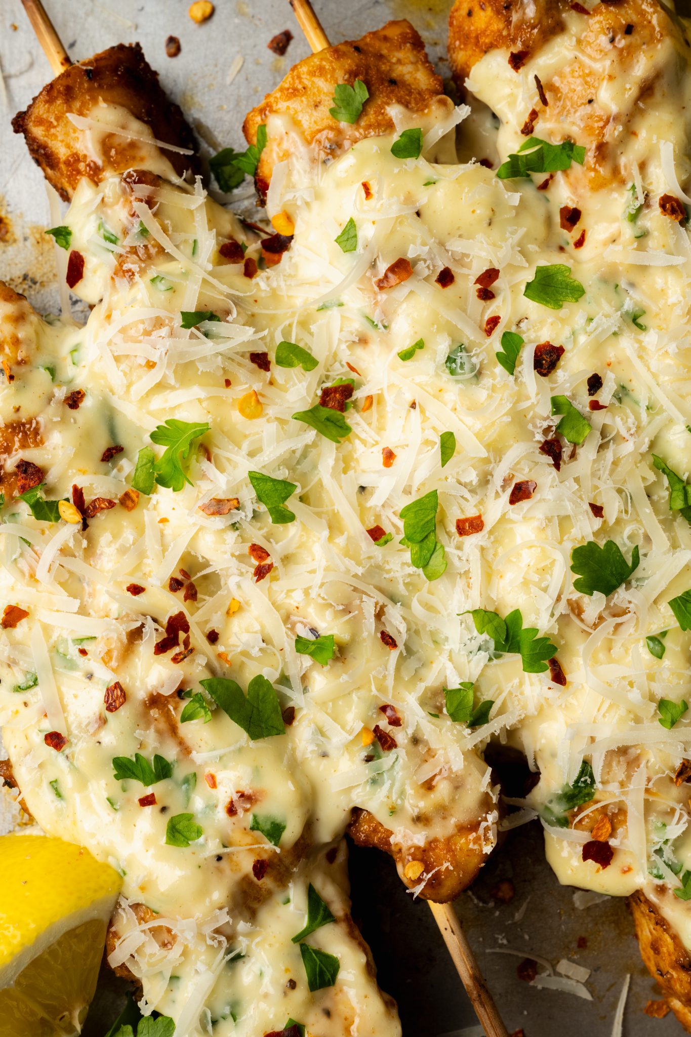 Chicken skewers fully brushed with garlic parmesan aioli and topped with shredded cheese, fresh parsley, and chili flakes on a metal sheet pan.