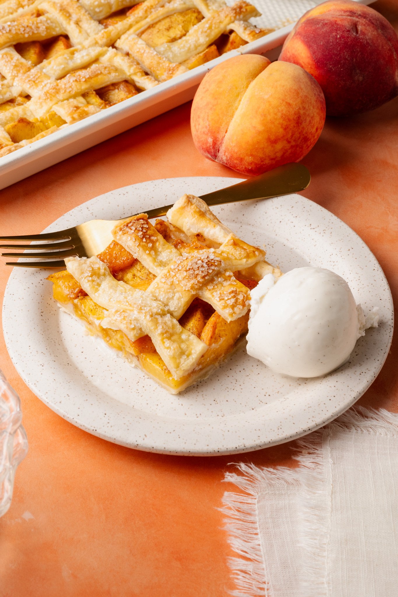 Side angle view of a plated slice of peach slab pie with a scoop of vanilla bean ice cream. The golden crust and vibrant peach filling are shown in detail with whole peaches in the background.