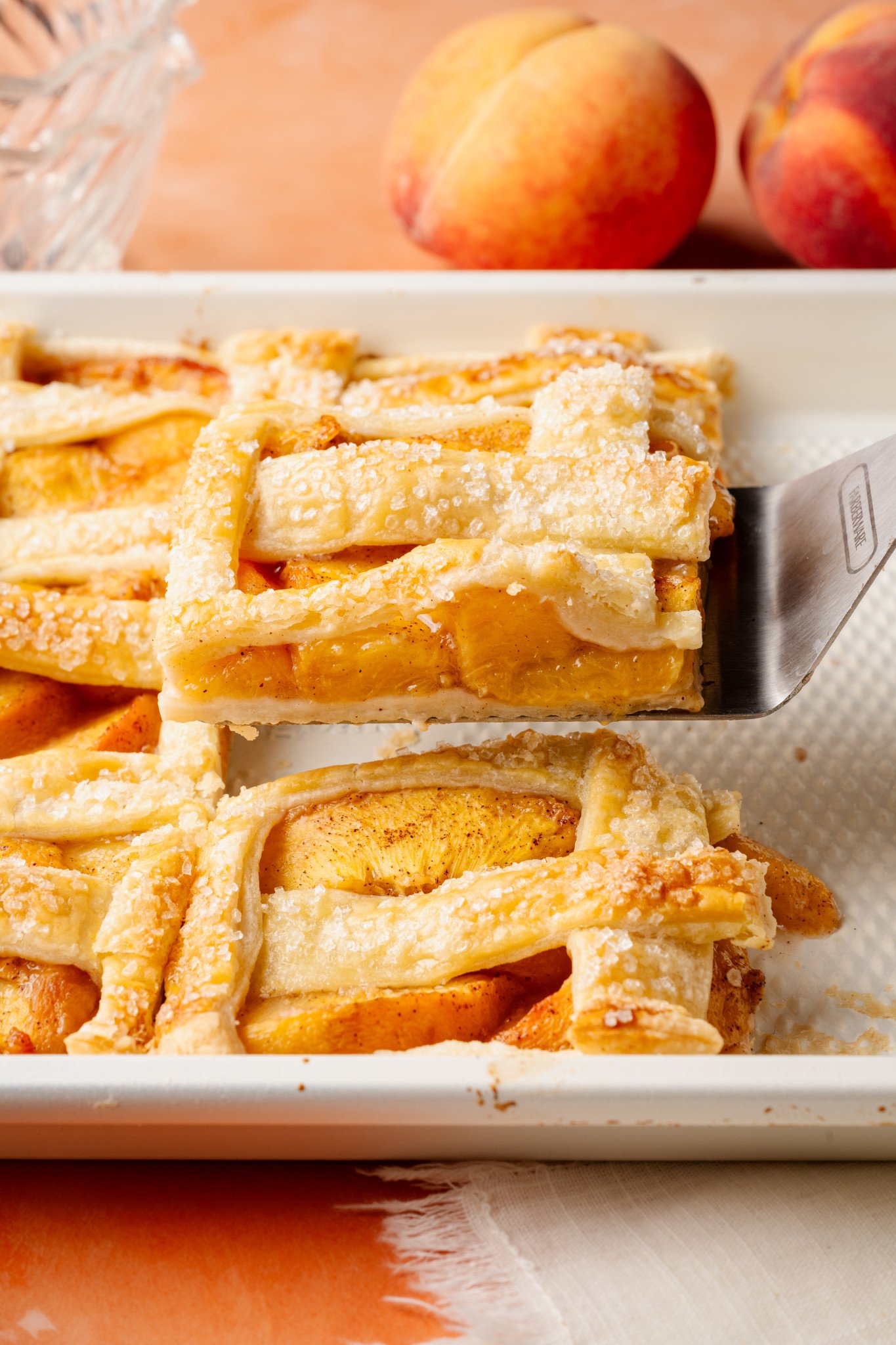 Close-up of a spatula lifting a square slice of peach slab pie from the baking dish. The crust is golden, flaky, and coated in coarse sugar, with tender peach slices visible underneath.
