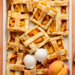 Top-down view of a peach slab pie that has been partially sliced and served, revealing its juicy filling. Three scoops of vanilla ice cream rest on top, and cinnamon sticks and peaches decorate the scene.