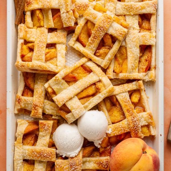 Top-down view of a peach slab pie that has been partially sliced and served, revealing its juicy filling. Three scoops of vanilla ice cream rest on top, and cinnamon sticks and peaches decorate the scene.