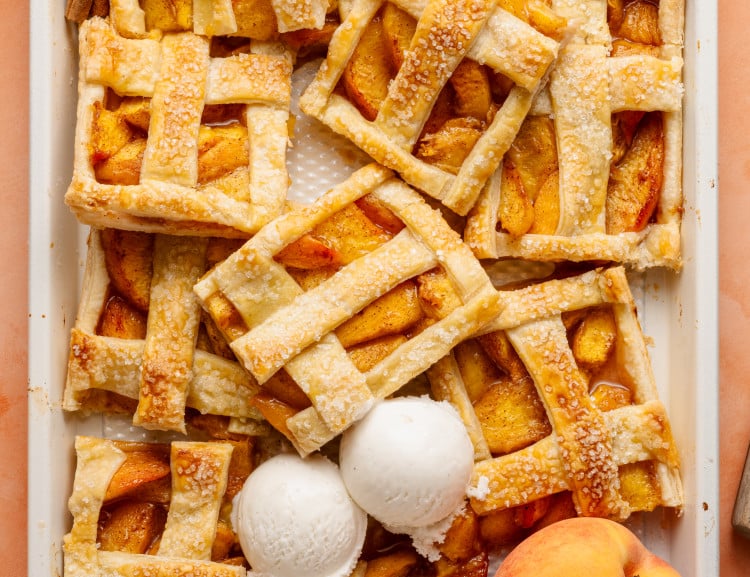 Top-down view of a peach slab pie that has been partially sliced and served, revealing its juicy filling. Three scoops of vanilla ice cream rest on top, and cinnamon sticks and peaches decorate the scene.