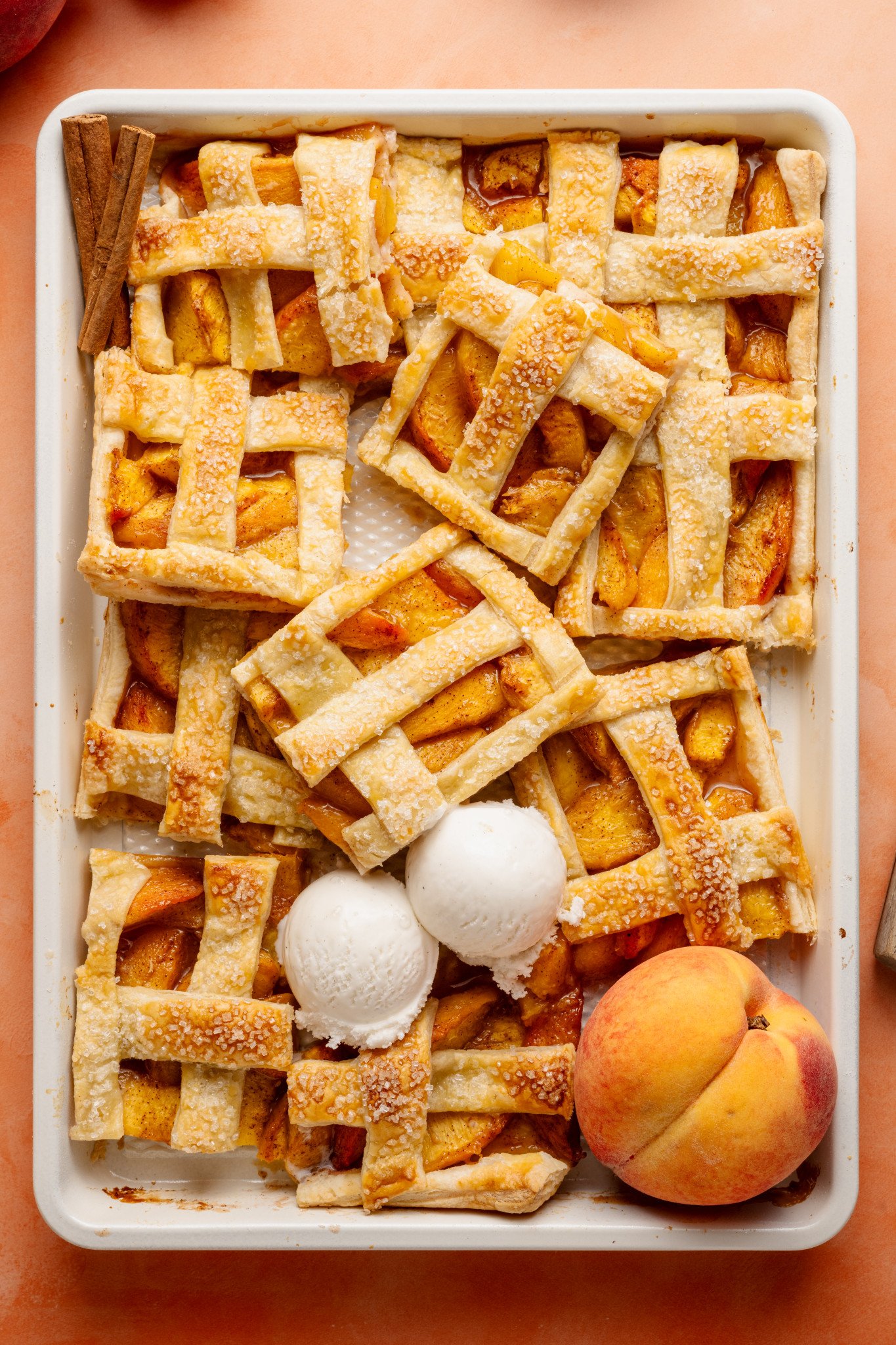 Top-down view of a peach slab pie that has been partially sliced and served, revealing its juicy filling. Three scoops of vanilla ice cream rest on top, and cinnamon sticks and peaches decorate the scene.