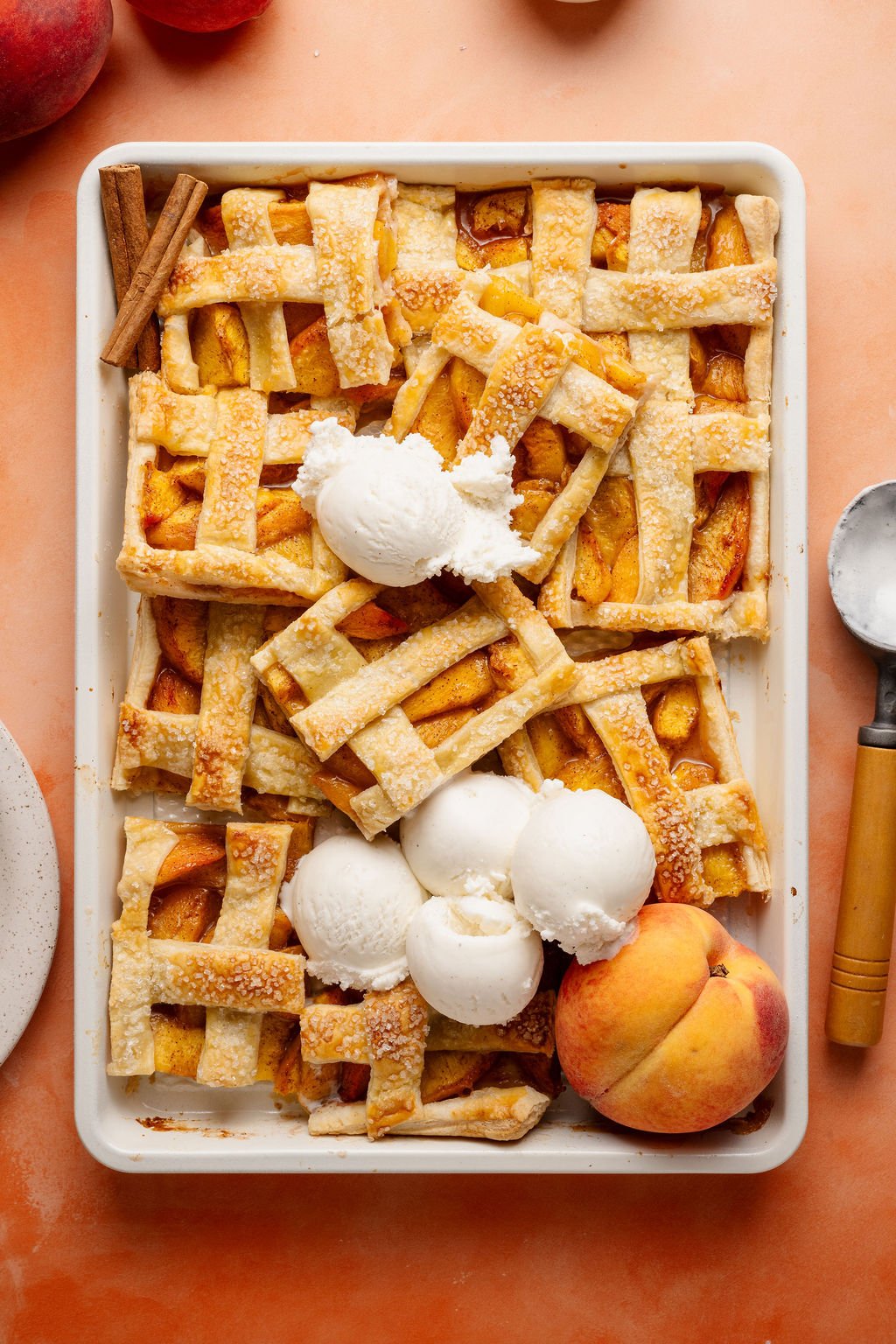 A tray of freshly baked peach slab pie bars with a golden, flaky lattice crust sprinkled with coarse sugar. Several bars are cut and slightly overlapping, topped with scoops of vanilla ice cream melting into the warm pie. A fresh peach and cinnamon sticks are placed beside the tray for garnish. The scene is set on a peach-colored surface with a metal ice cream scoop nearby.