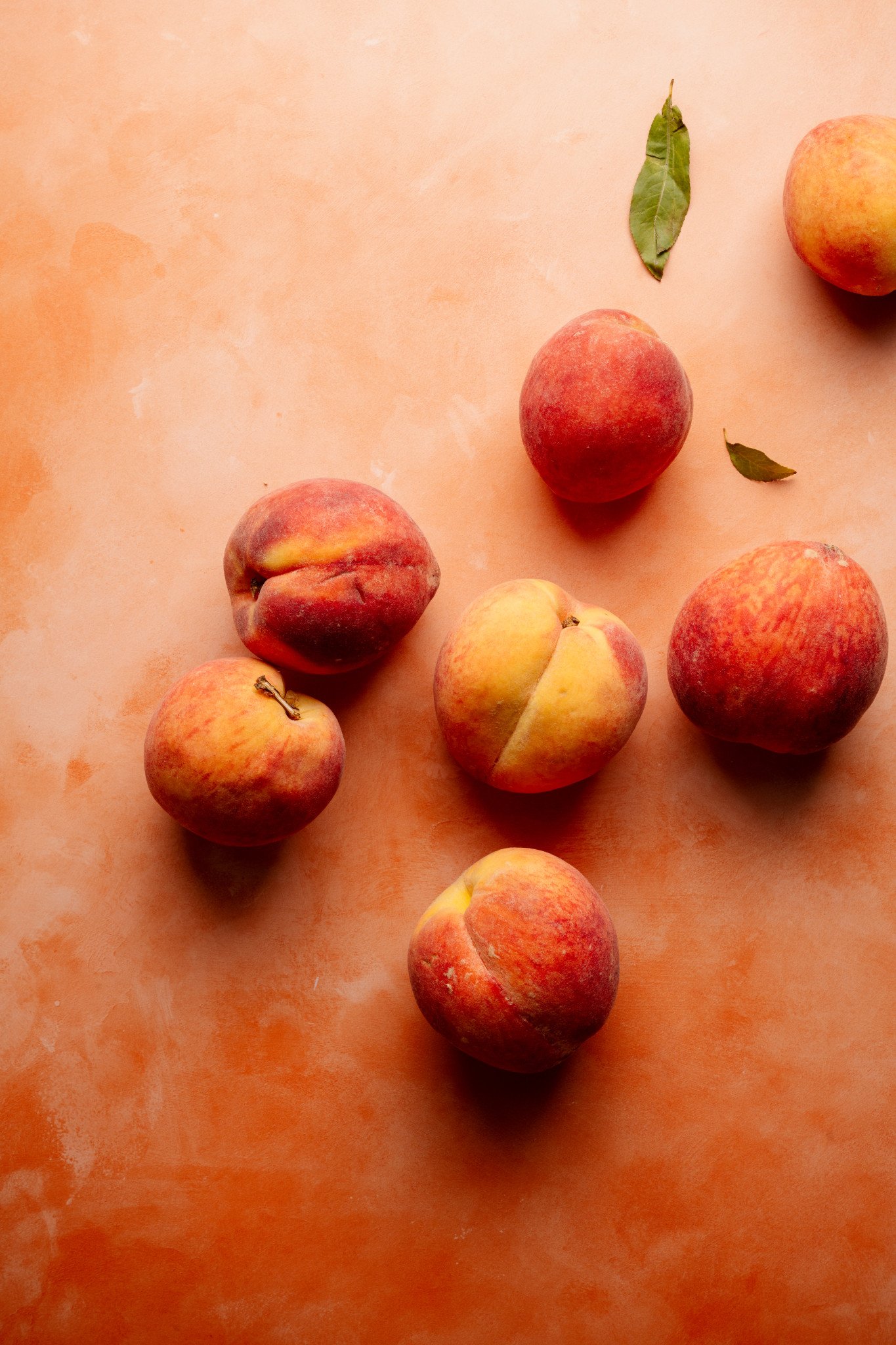 Flat lay of fresh whole peaches scattered on a peach-colored background with a few green leaves. The peaches vary slightly in shape and color, adding visual texture.