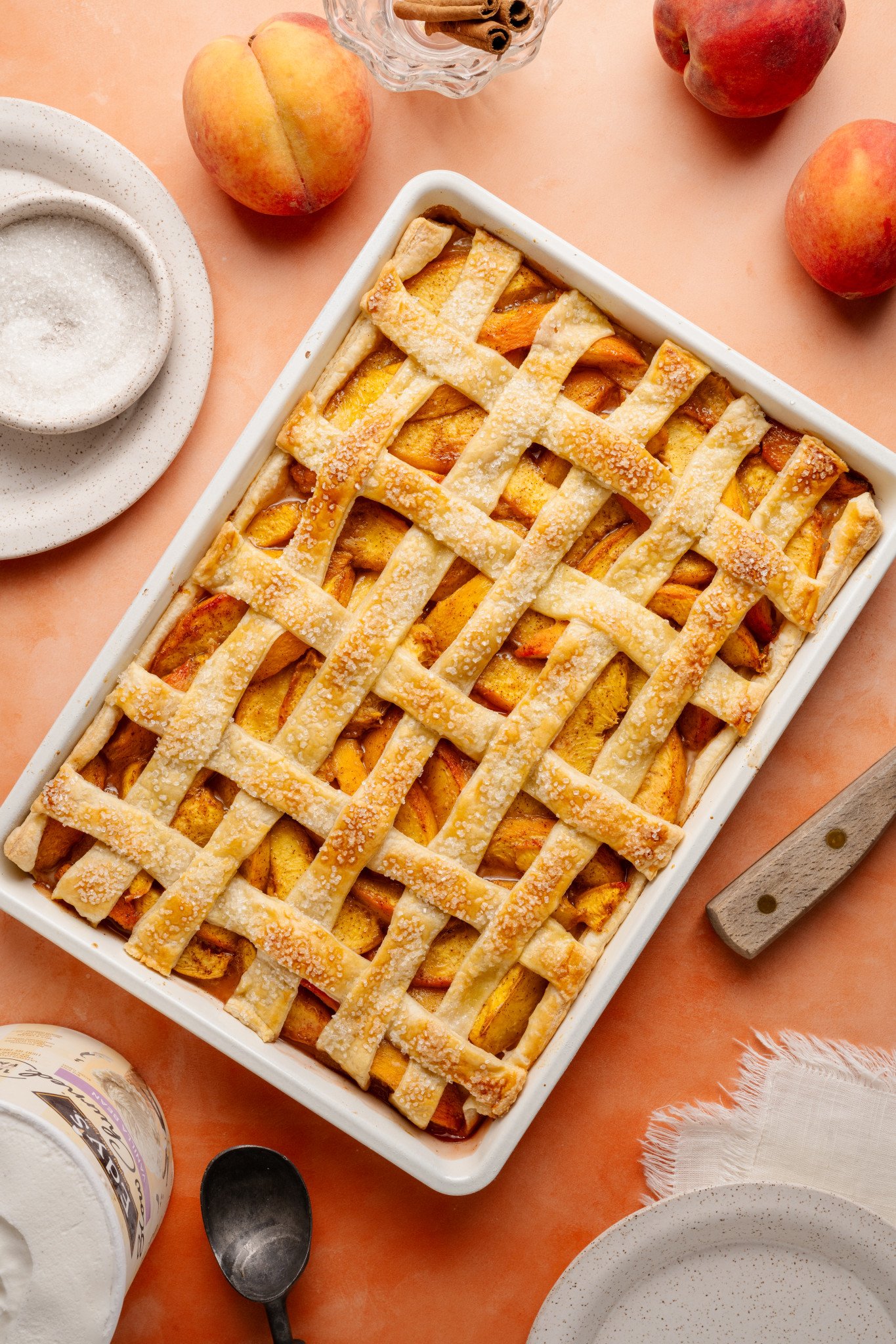 Overhead view of a golden-brown peach slab pie in a white rectangular baking dish. The lattice crust is sprinkled with coarse sugar, and fresh peaches, cinnamon sticks, and plates surround the dish on a peach-colored background.