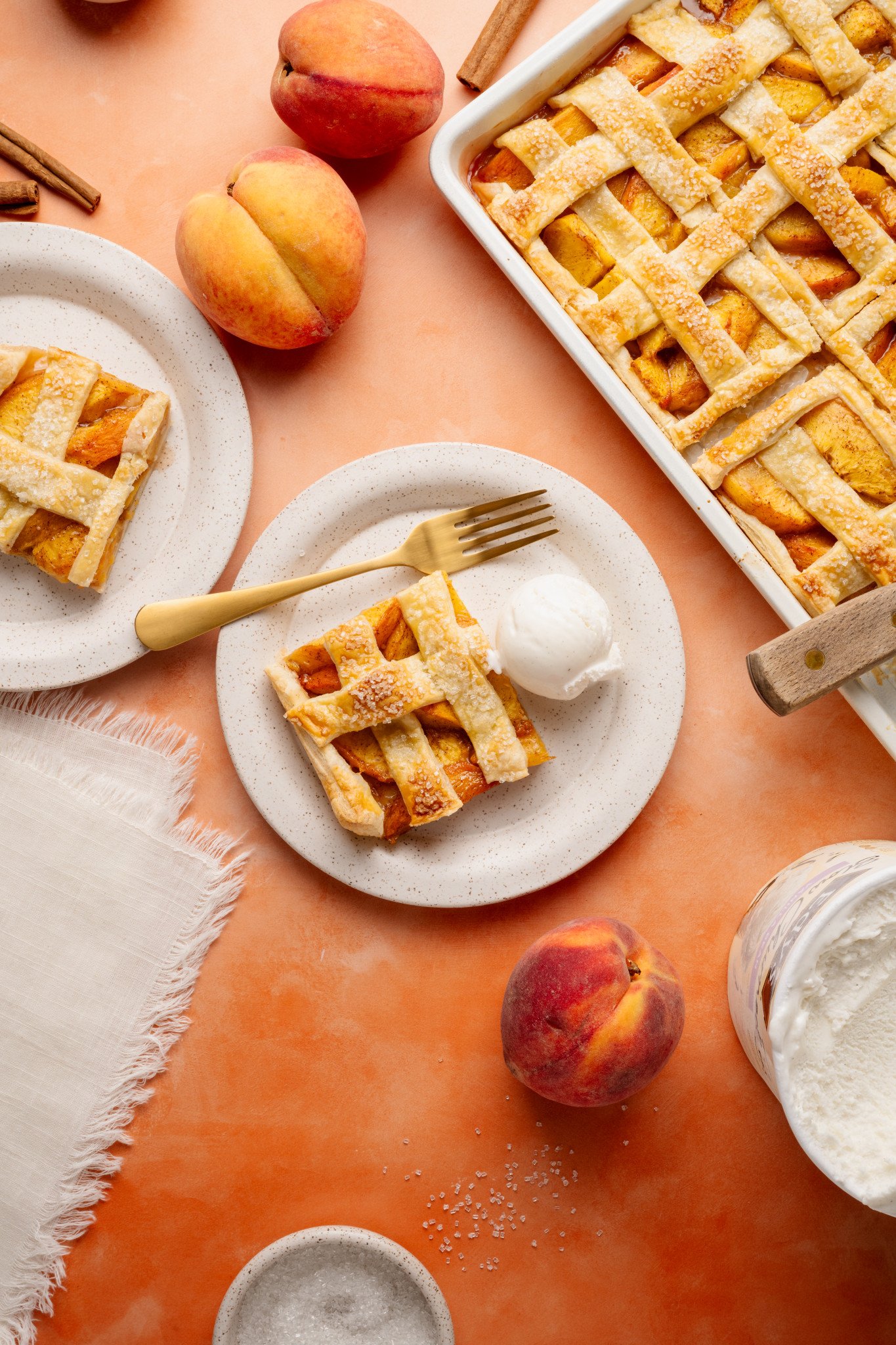 Two slices of peach slab pie on speckled plates, one served with a scoop of vanilla ice cream and a gold fork. The full pie is partially visible in the corner, with fresh peaches and cinnamon sticks scattered around.