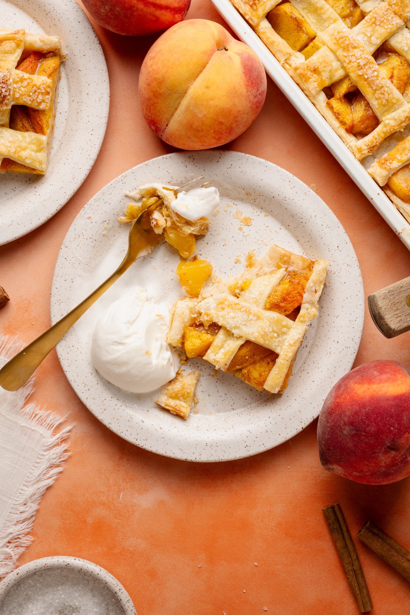A serving of peach slab pie mid-bite, with a fork holding a piece of flaky crust, juicy peaches, and a bit of ice cream. The remaining pie and whole peaches are arranged nearby on the warm-toned background.