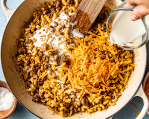 Pouring cream and adding the shredded cheese to hamburger macaroni mixture in pot.
