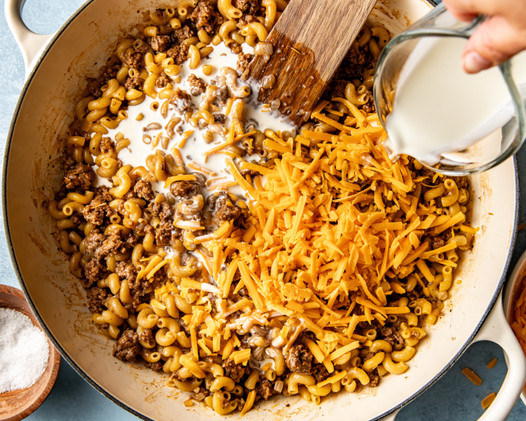 Pouring cream and adding the shredded cheese to hamburger macaroni mixture in pot.