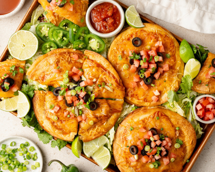 A colorful serving tray with fully assembled Mexican pizzas sliced and garnished with diced tomatoes, black olives, green onions, and fresh cilantro. Lime wedges, jalapeños, salsa, and shredded lettuce surround the pizzas for serving.