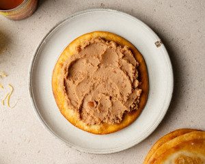 A fried tortilla placed on a white plate is topped with a smooth layer of refried beans, evenly spread across the surface.