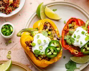 Overhead view of colorful quinoa stuffed bell peppers on a ceramic plate, garnished with sour cream, sliced jalapeños, green onions, and cilantro, with lime wedges on the side.