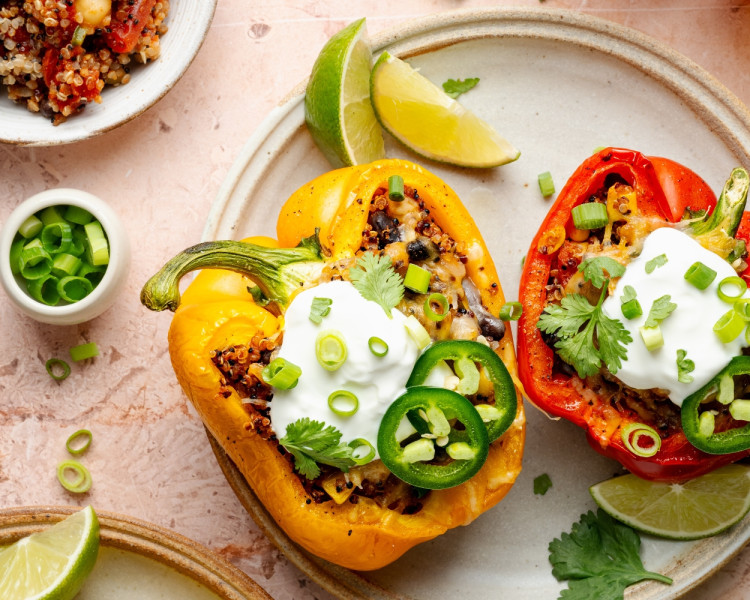 Overhead view of colorful quinoa stuffed bell peppers on a ceramic plate, garnished with sour cream, sliced jalapeños, green onions, and cilantro, with lime wedges on the side.