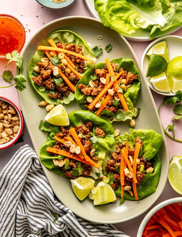 Four Thai chicken lettuce wraps on large oval plate garnished with pickled carrots, peanuts and lime wedge.
