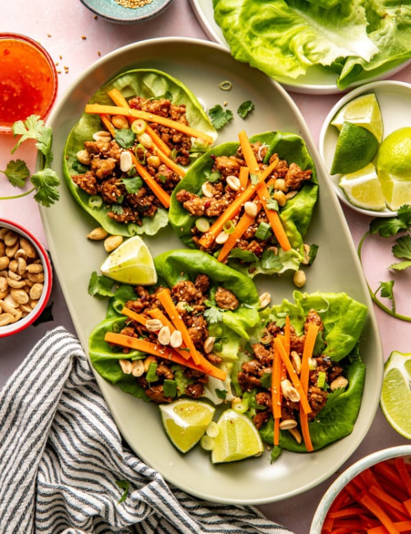 Four Thai chicken lettuce wraps on large oval plate garnished with pickled carrots, peanuts and lime wedge.