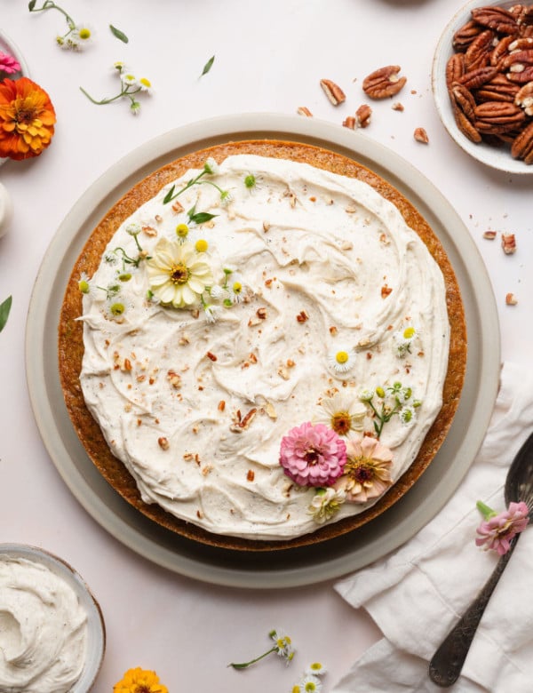 Overhead view of a whole zucchini cake on a plate, generously frosted with vanilla frosting and decorated with chopped pecans and pastel edible flowers.