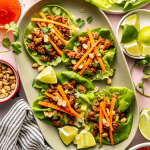 Four Thai chicken lettuce wraps on large oval plate garnished with pickled carrots, peanuts and lime wedge.