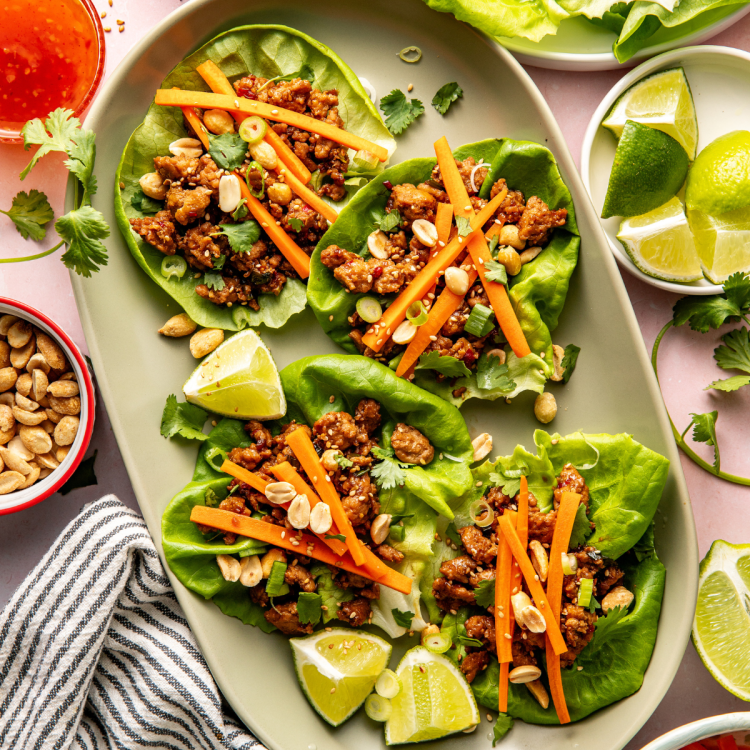 Four Thai chicken lettuce wraps on large oval plate garnished with pickled carrots, peanuts and lime wedge.