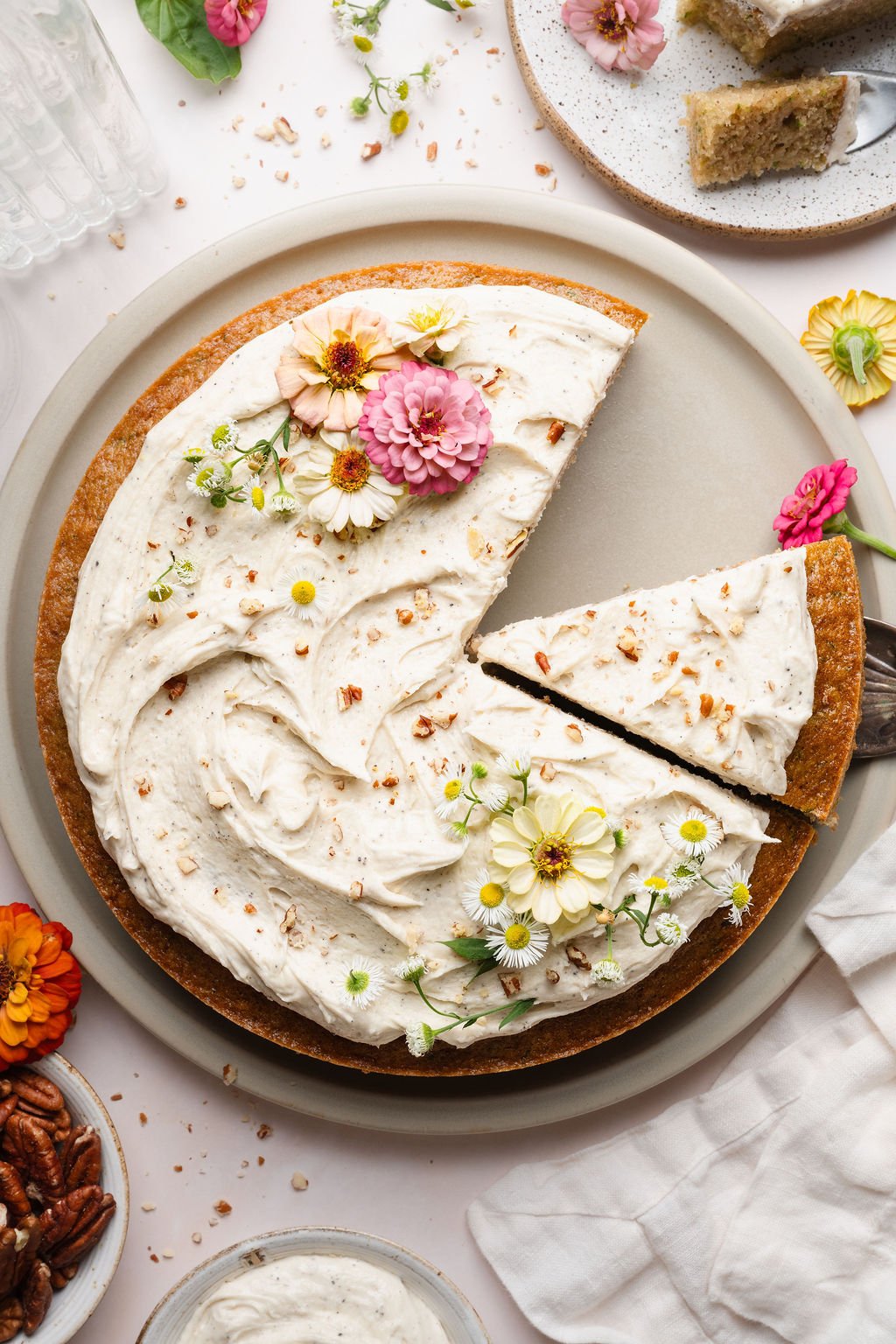 A round zucchini cake topped with fluffy vanilla frosting, garnished with edible flowers and chopped pecans. A slice has been cut out and placed on a cake server, revealing the moist interior.