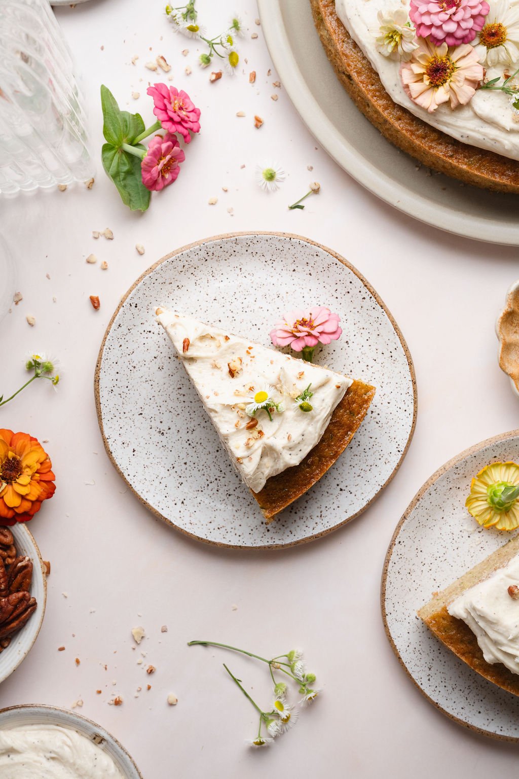 A single slice of zucchini cake with creamy frosting and a sprinkle of pecans on a speckled ceramic plate, surrounded by zinnias and chamomile flowers.
