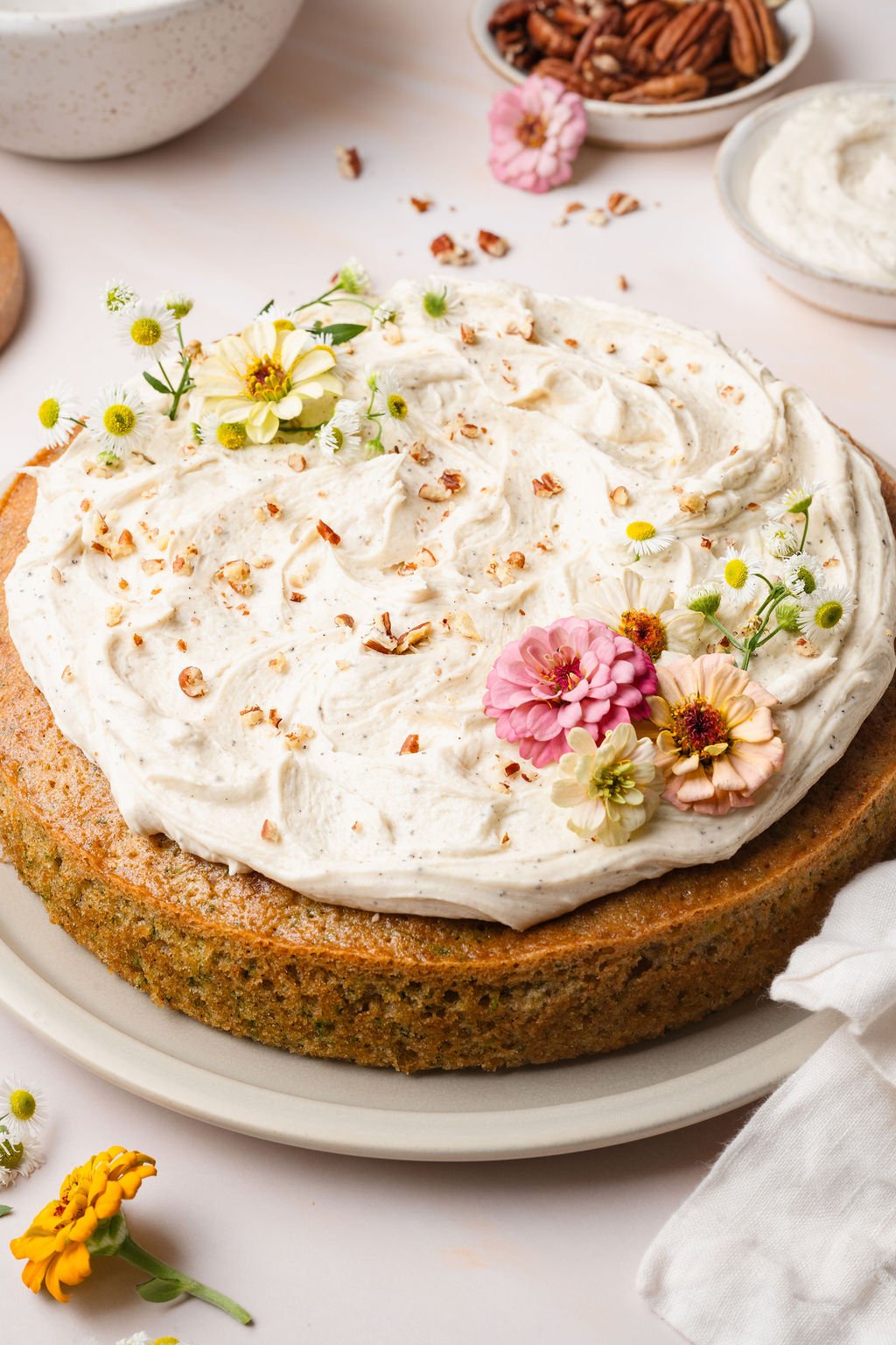 Side view of a whole zucchini cake with thick swirls of vanilla frosting on top, garnished with colorful zinnias and daisies.