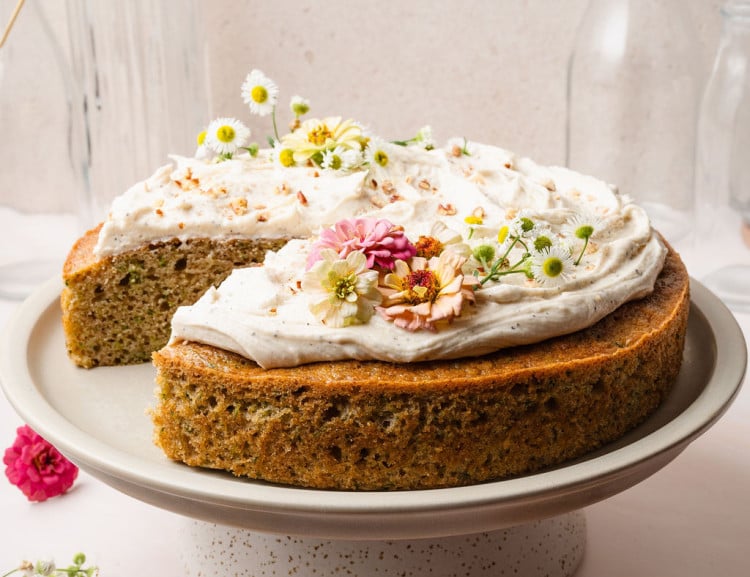 A zucchini cake with a missing slice, showing its tender green-flecked interior. The cake is topped with vanilla frosting and fresh flowers on a pedestal.