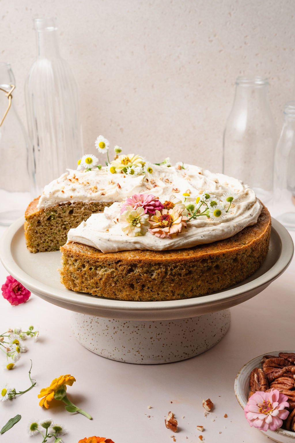 A zucchini cake with a missing slice, showing its tender green-flecked interior. The cake is topped with vanilla frosting and fresh flowers on a pedestal.