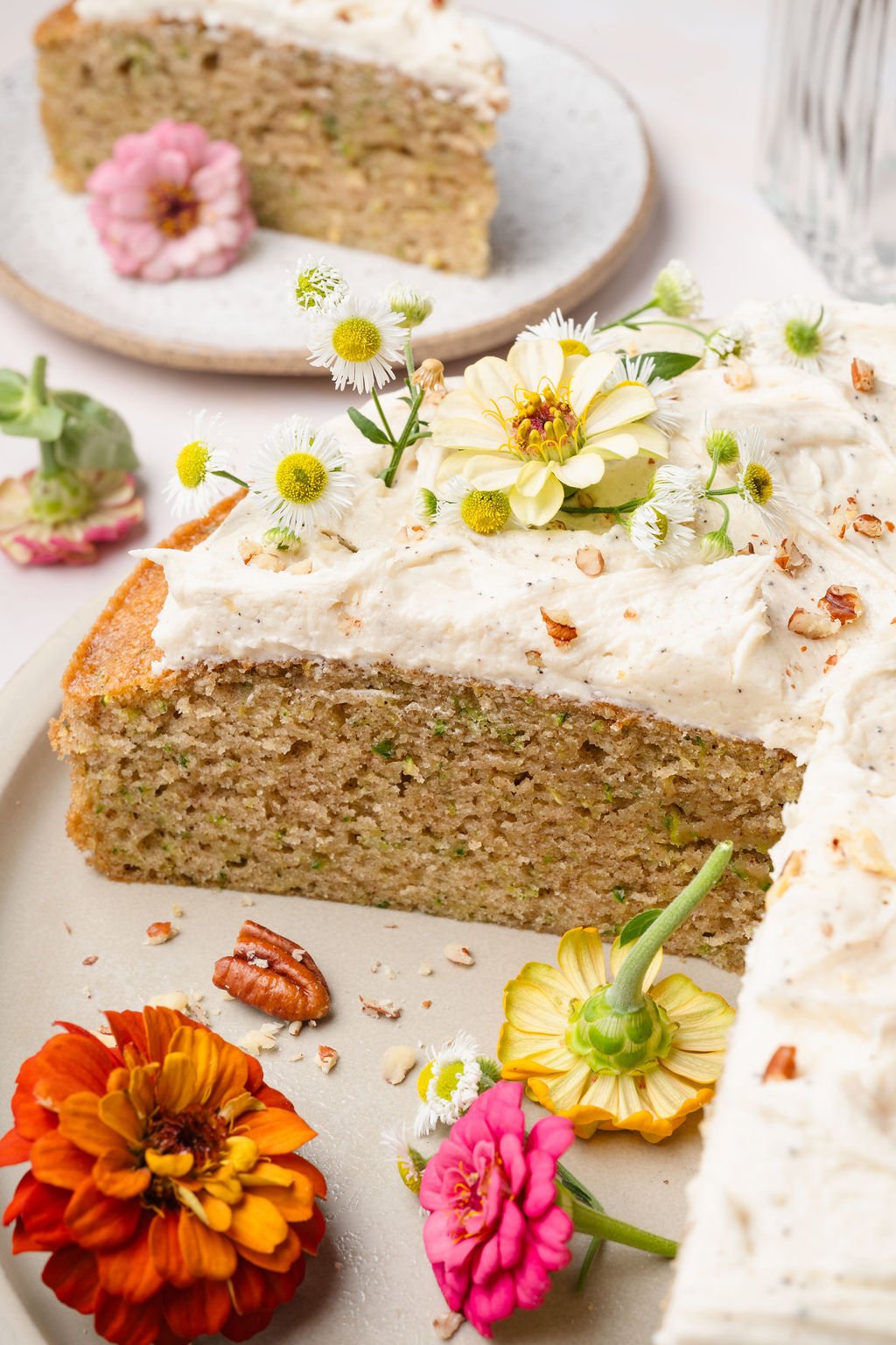 Close-up of a sliced zucchini cake showing its tender, speckled crumb. The cake is topped with thick vanilla frosting, chopped pecans, and fresh edible flowers.