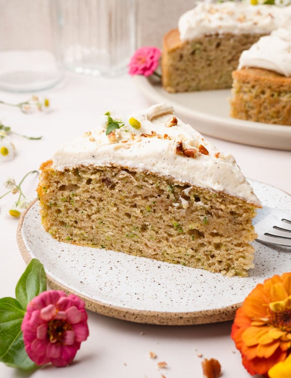 A plated slice of zucchini cake topped with frosting and chopped nuts, with the full cake visible in the background. Surrounded by bright zinnias.