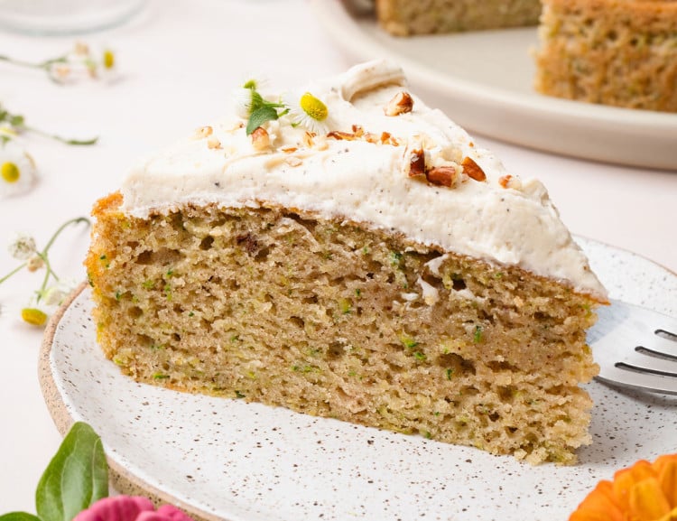 A plated slice of zucchini cake topped with frosting and chopped nuts, with the full cake visible in the background. Surrounded by bright zinnias.