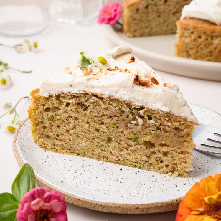 A plated slice of zucchini cake topped with frosting and chopped nuts, with the full cake visible in the background. Surrounded by bright zinnias.