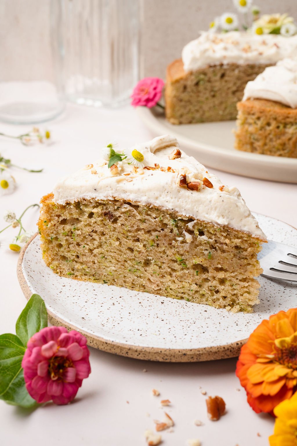 A plated slice of zucchini cake topped with frosting and chopped nuts, with the full cake visible in the background. Surrounded by bright zinnias.