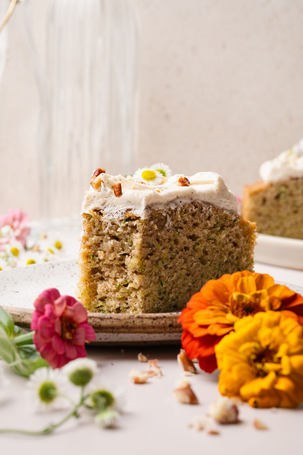 A thick slice of zucchini cake with speckled frosting and chopped pecans on a ceramic plate. Colorful zinnias and daisies add a playful touch to the scene.