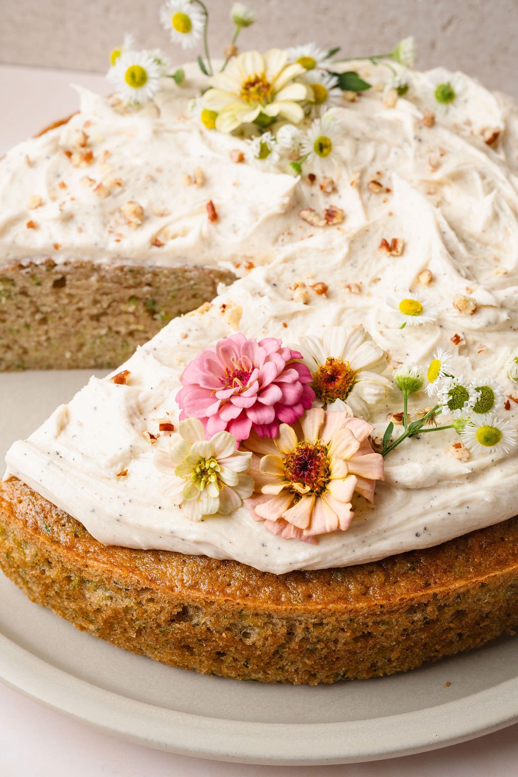 Close-up of a zucchini cake topped with creamy frosting, chopped pecans, and pastel edible flowers. A slice has been removed, revealing the moist green-flecked interior.