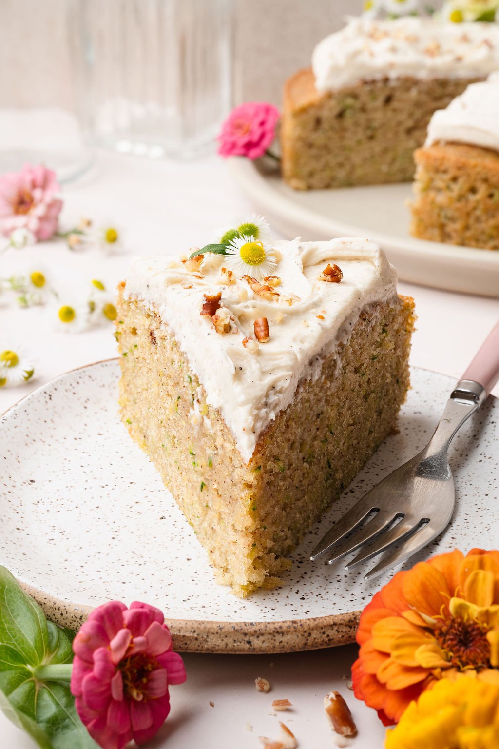 A triangular slice of zucchini cake on a speckled plate, topped with creamy frosting, chopped pecans, and a tiny daisy. Bright zinnias surround the plate.
