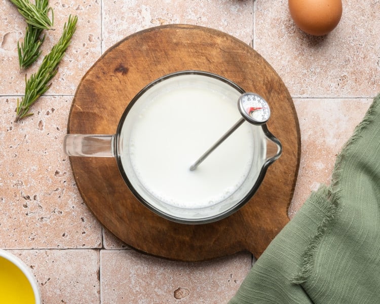 A glass measuring cup filled with warm milk on a wooden board with a thermometer inside, surrounded by fresh rosemary and an egg.