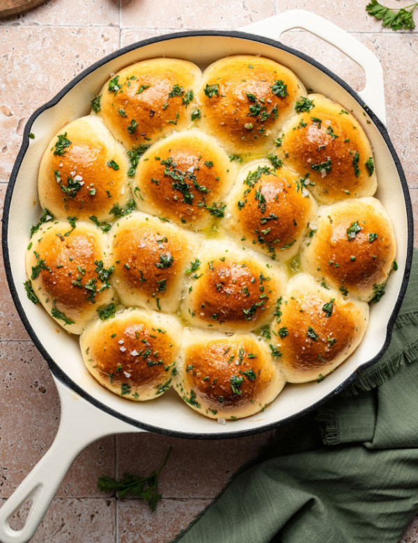 Golden brown garlic rosemary pull-apart rolls baked in a round white skillet, brushed with melted butter and sprinkled with fresh herbs.