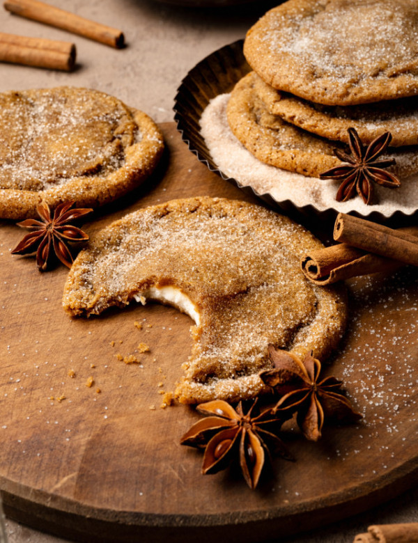 Pumpkin cheesecake cookie with bite taken out of it on wood board with stack of three more cookies in cinnamon sugar bowl behind it.