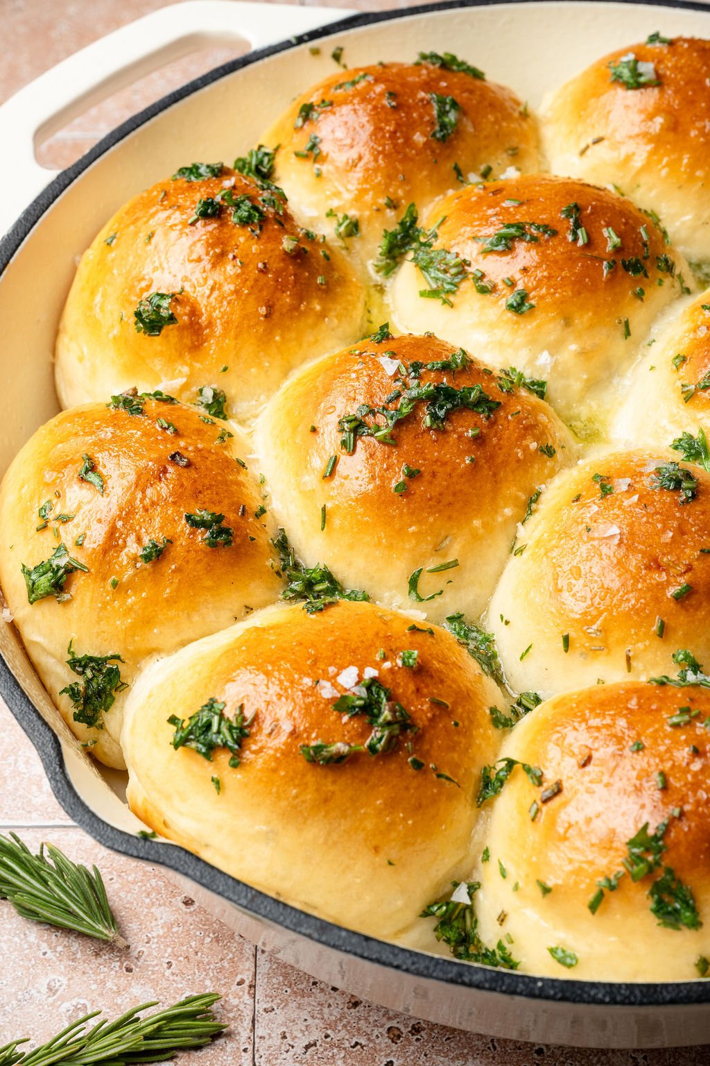 Freshly baked garlic rosemary rolls in a white skillet, topped with melted butter, herbs, and sea salt.