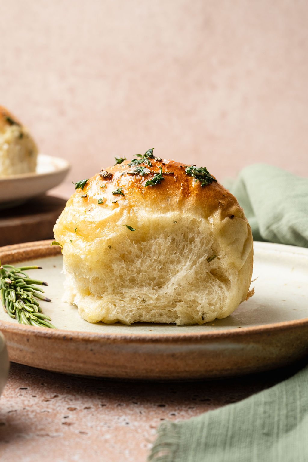 Single garlic rosemary roll on a ceramic plate, showing its fluffy inside and shiny golden top with herbs.
