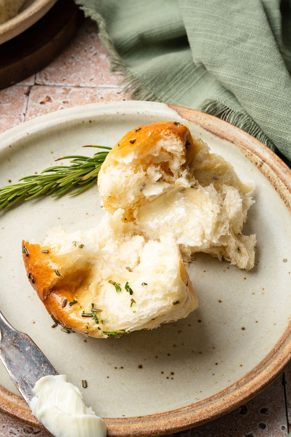 Torn garlic rosemary roll on a plate with melted butter inside, next to a butter knife and fresh rosemary sprig.