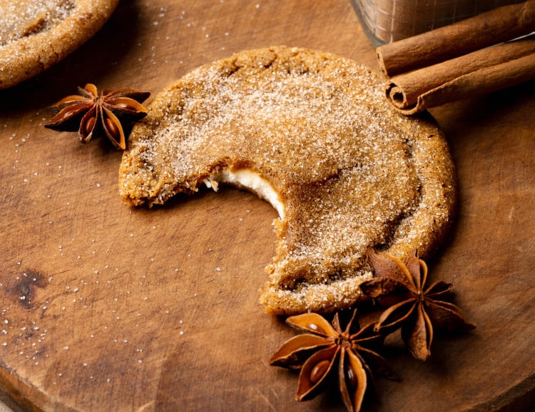 Pumpkin cheesecake cookie with bite taken out of it on wood cutting board with cinnamon cloves around it.