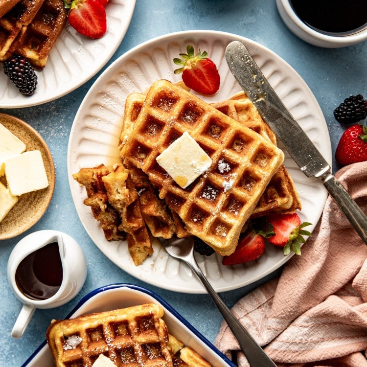 Plate of buttermilk waffles topped with butter and syrup with fork cutting into the waffles and butterknife on the side of the plate.