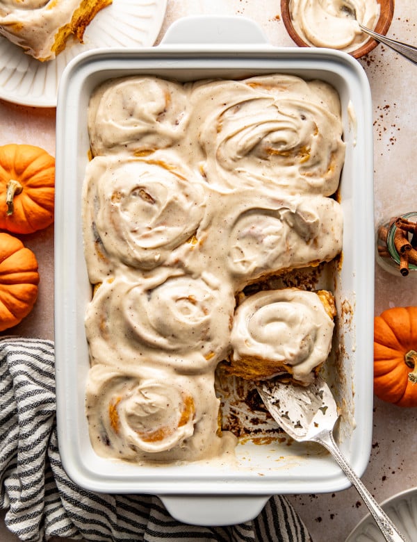 Seven frosted pumpkin cinnamon rolls in 9x13 baking pan with spatula removing one roll.