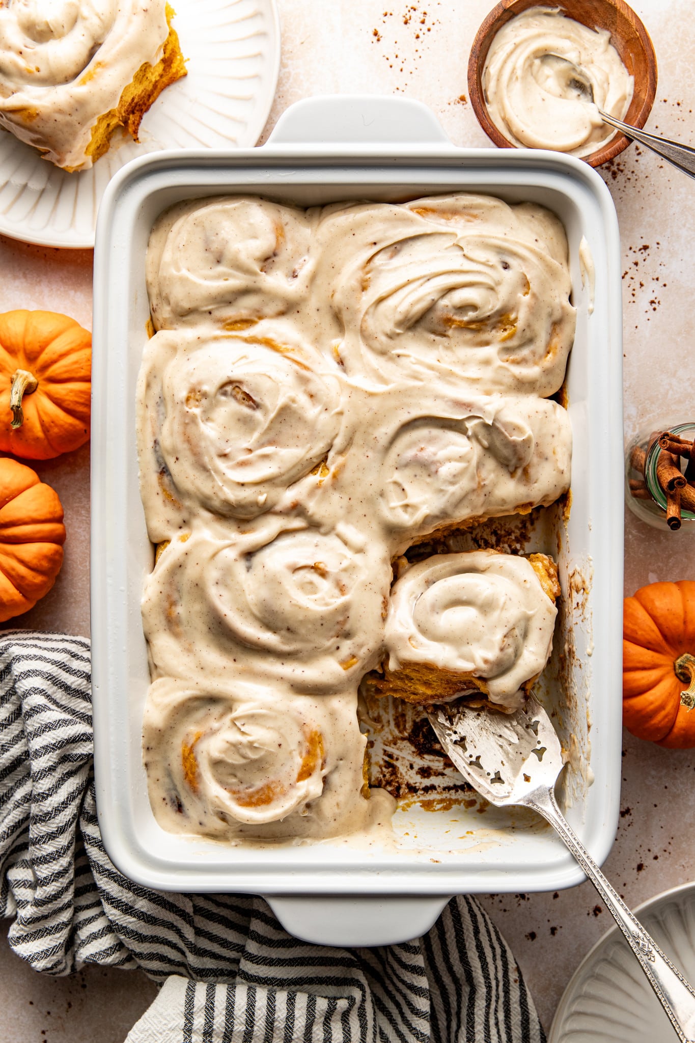 Seven frosted pumpkin cinnamon rolls in 9x13 baking pan with spatula removing one roll.