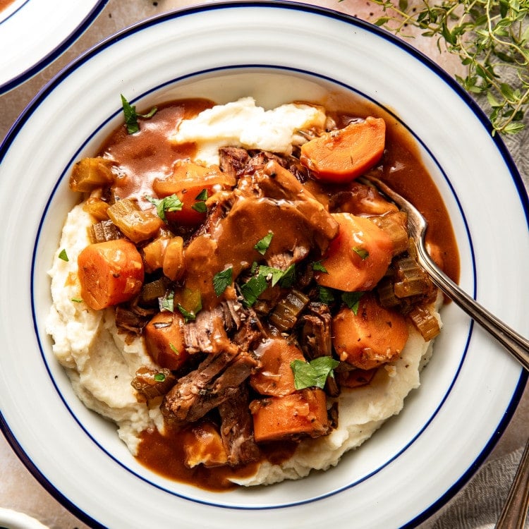 Slow cooker pot roast served over mashed potatoes in a white serving bowl with fork.