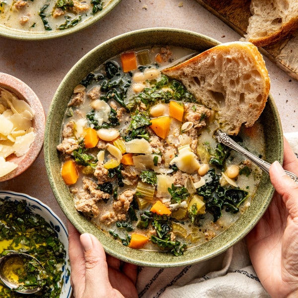 Sausage, kale and white bean soup in bowl with a side of crusty bread and spoon in bowl.